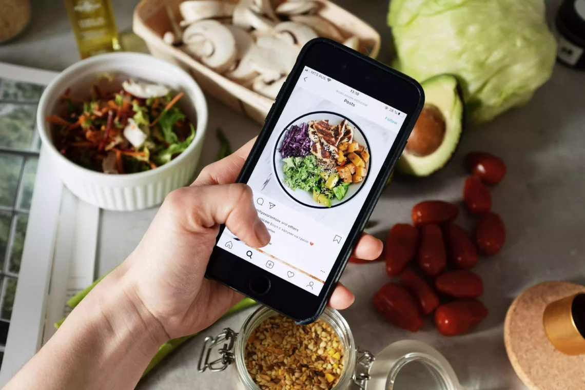 algorithme instagram . Photo d'une main tenant un smartphone avec l'application Instagram ouverte. On voit un post Instagram avec la photo d'une assiette colorée et appétissante. En arrière plan, on voit une table avec de nombreux ingrédients comme des légumes.
