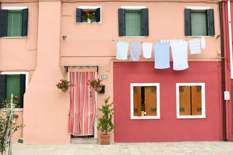 façade de maison colorée, dans les tons roses et terracotta, avec les volets fermés, un rideau devant la porte, et du linge suspendu à une corde à linge le long des fenêtres du haut